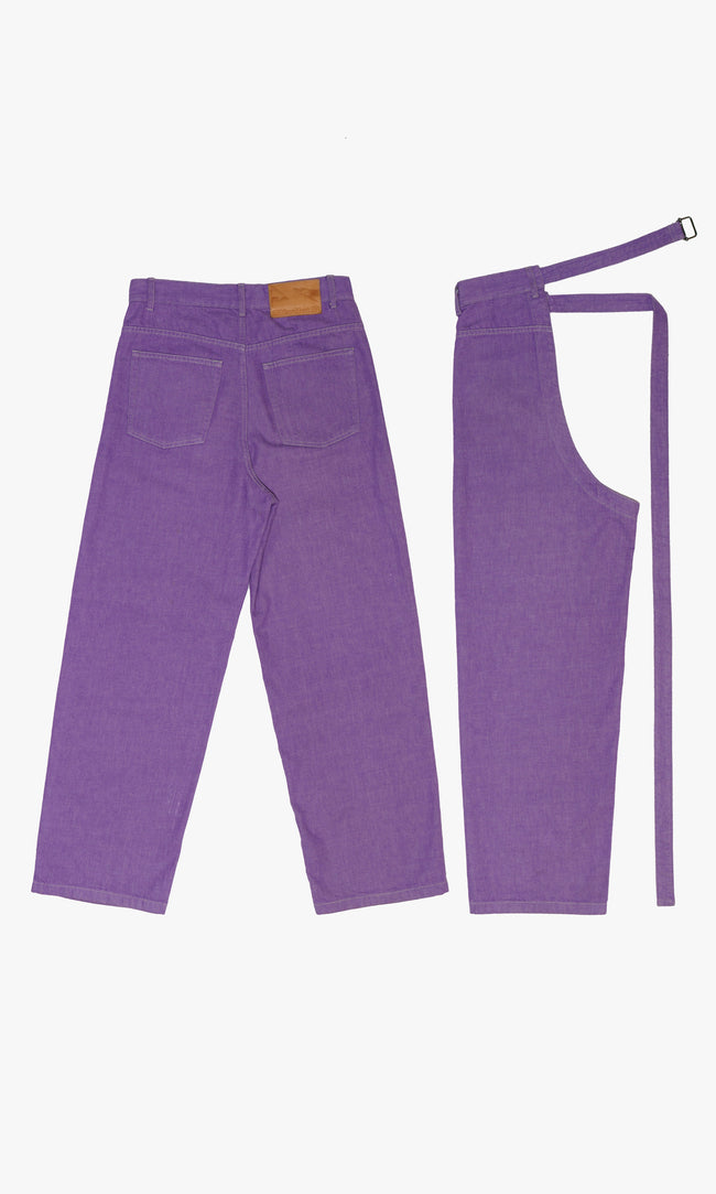 SIEGENTHALER | Back view of the modular jeans with chaps in purple denim. Featuring double patch pockets and a tan leather logo patch, the silhouette contrasts classic denim workwear codes with a sculptural layered cut.