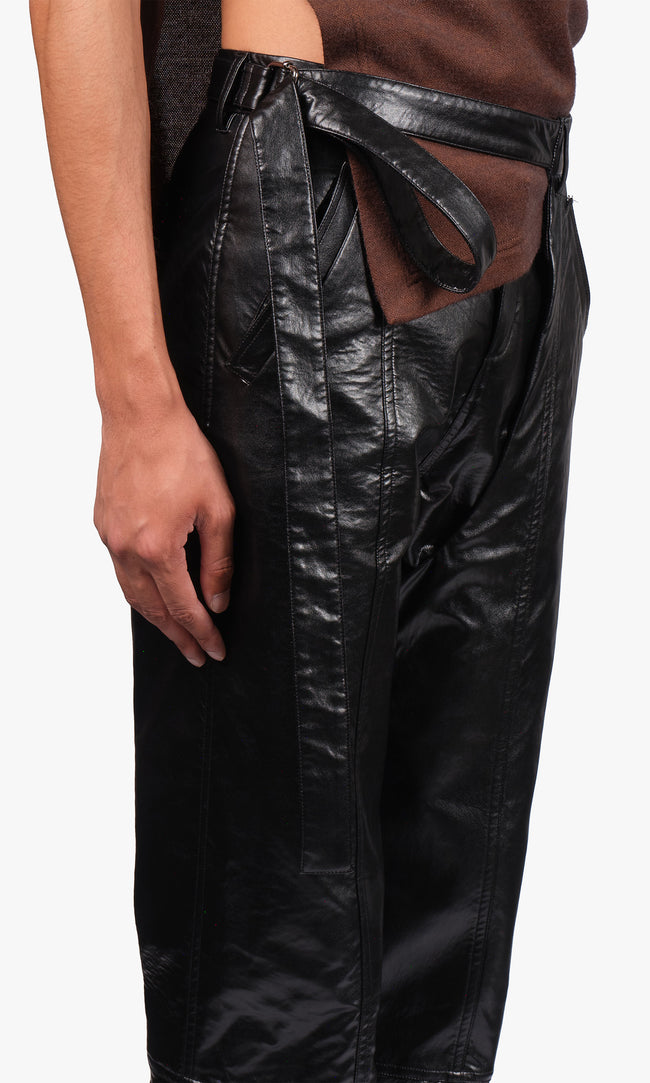 SIEGENTHALER | Side detail view of the black faux leather pants with chaps, showcasing the layered construction and articulated curved seaming. A wrapped belt element extends from the waistband, adding a sculptural and adjustable feature to the sleek silhouette.