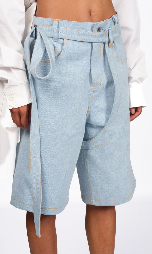 SIEGENTHALER | Front detail view of the light blue denim shorts, focusing on the architectural seam lines, angled waistband with integrated belt, and oversized curved panel construction that accentuates the leg movement.