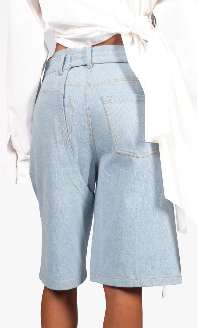 SIEGENTHALER | Back view of the shorts in light blue denim, showcasing curved panel construction, contrast topstitching, and raw edge strap detail extending from the hem.