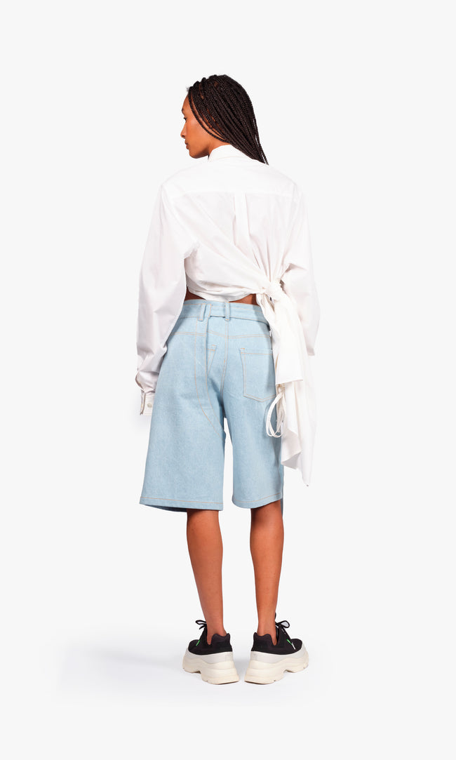 SIEGENTHALER | Back view of the light denim shorts, featuring contrasted stitching and a sculpted rear yoke. Modular belt system loops through the waistband while curved panel lines hint at the integrated chaps construction.