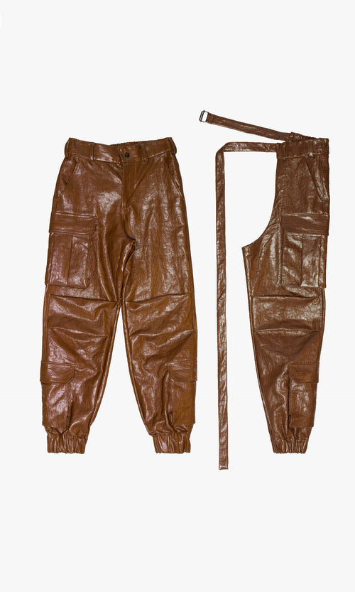 SIEGENTHALER | Front view of the brown faux leather cargo pants with chaps, showcasing modular construction with elongated wraparound panel and extended belt. Utility flap pockets, elasticated cuffs, and a structured shine give the silhouette a sculptural streetwear edge.