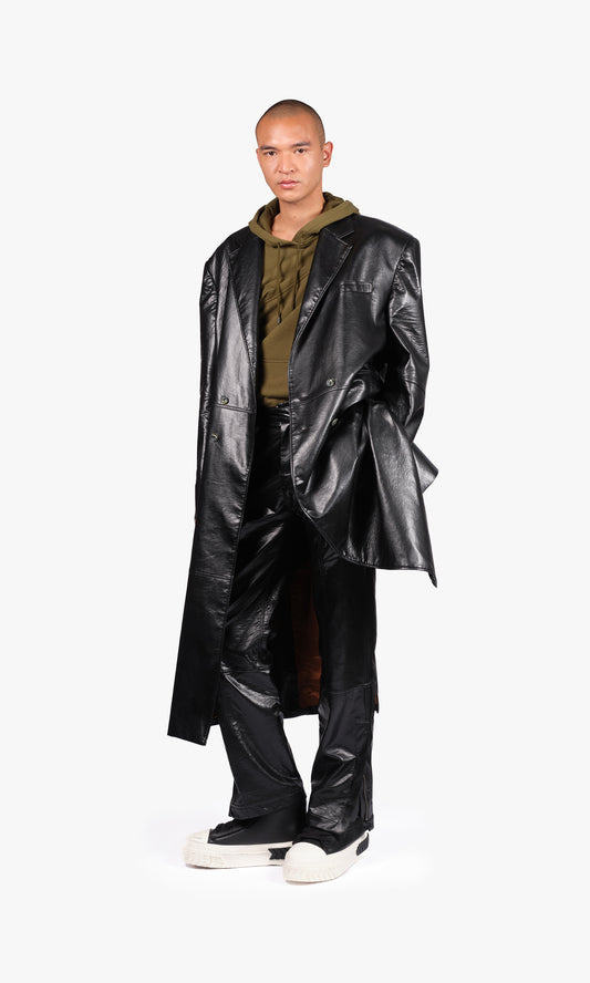 SIEGENTHALER | front view of the long coat in black faux leather. Oversized silhouette with wide sleeves, dropped shoulders, and a long slit at the back. Tailored in glossy faux leather with structured volume and full lining.