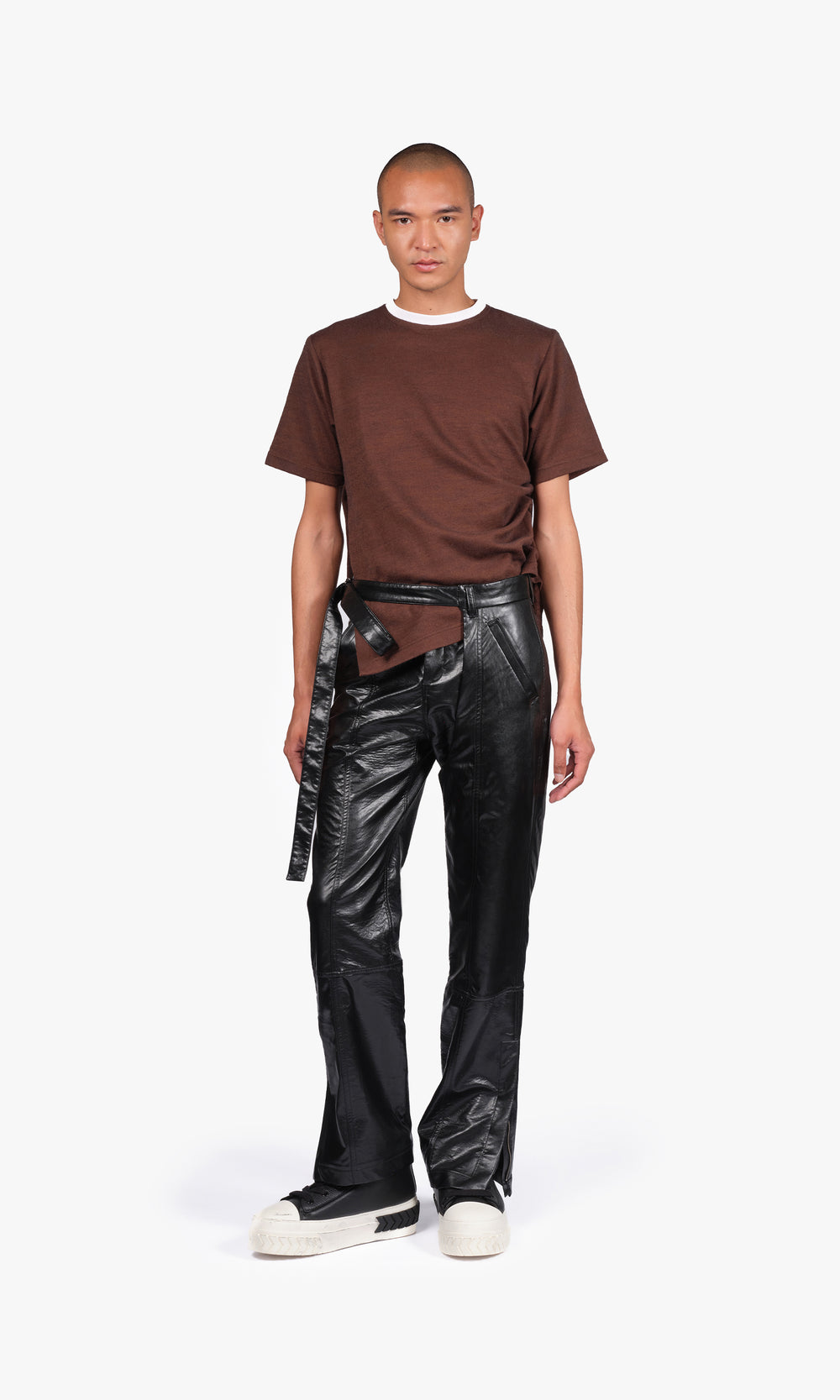 SIEGENTHALER | Front view of the black faux leather pants with chaps, constructed with articulated paneling and a modular silhouette. The trousers feature detachable chaps, a signature curved seam design, and a long belt tie extending from the waistband. The glossy finish enhances the garment’s sculptural shape and fluidity.
