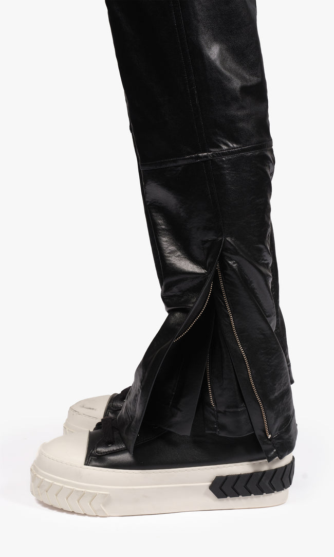 SIEGENTHALER | Hem detail view of the black faux leather pants with chaps, showing the lower-leg zippered opening and layered panel construction. The exaggerated split adds dynamic volume and reveals a glimpse of the inner structure.